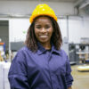 Cheerful African American female factory employee in hardhat and overall standing on plant floor, looking at camera and smiling. Front view, medium shot. Women in industry concept